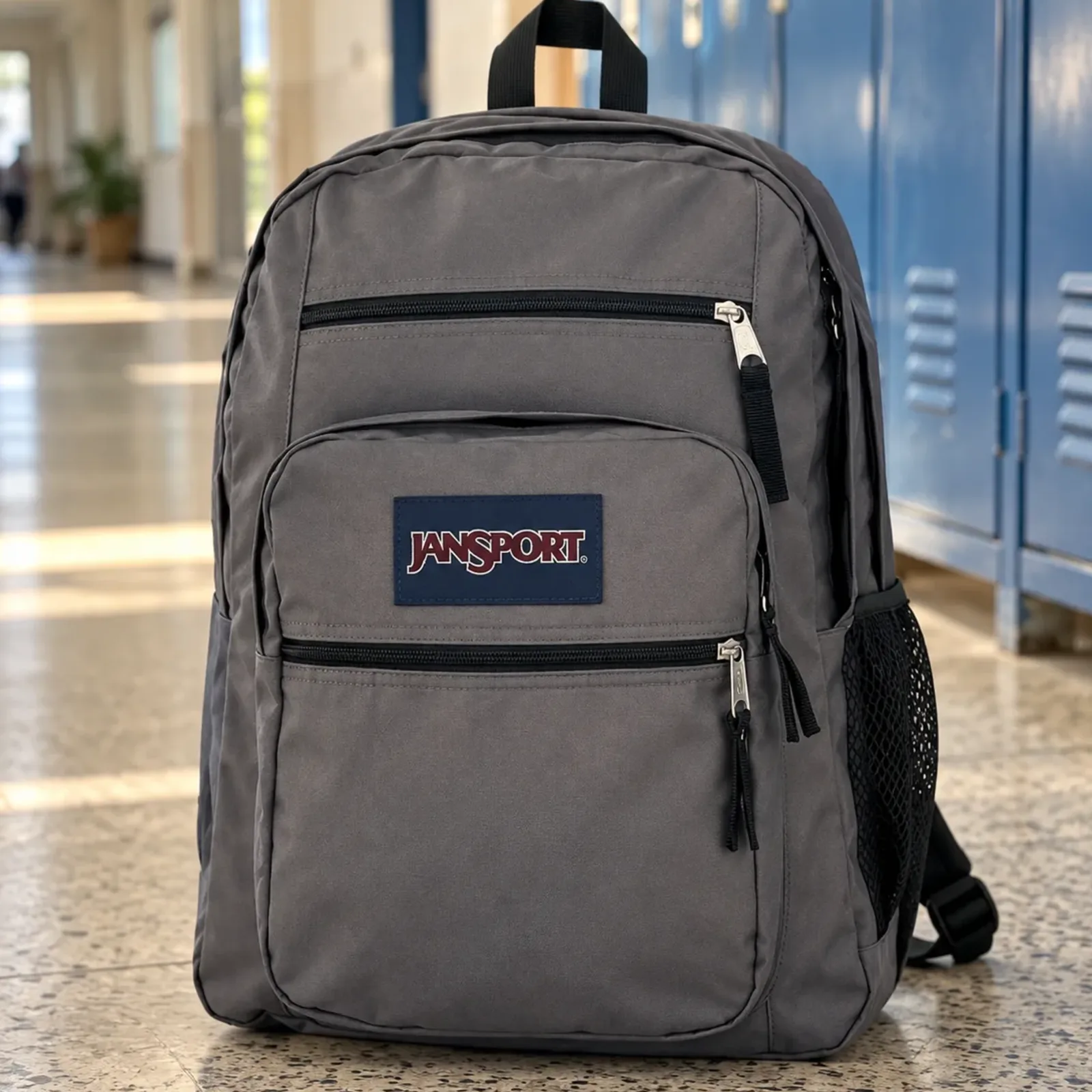 Mochila Big Student cinza grafite em pé no piso de um corredor de escola, com os armários azuis fora de foco