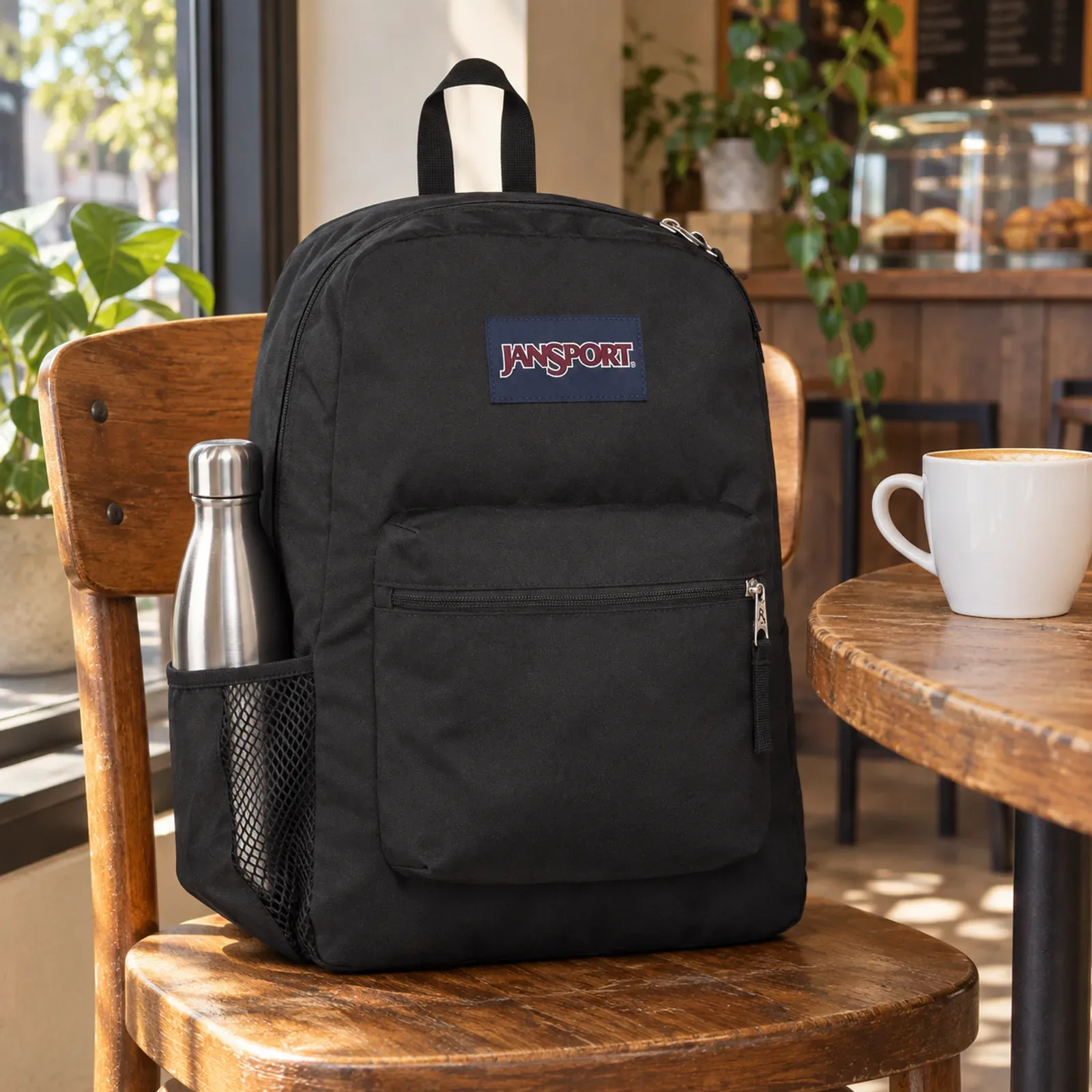 Mochila Cross Town preta apoiada no assento de uma cadeira de madeira, perto da janela de um café, garrafa de inox no bolso de tela